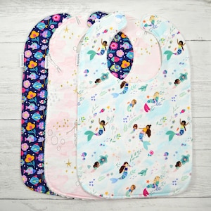 May include: Three baby bibs with snap closures. The first bib features a dark blue background with colorful fish and flowers. The second bib is light pink with gold stars and mermaid outlines. The third bib is white with mermaids and sea life.