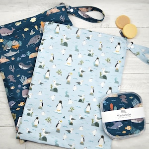 May include: Two reusable cloth bags with marine life patterns. One bag is navy blue with whales, fish, and coral. The other is light blue with penguins and seagulls. Also included are two round soap bars and a set of washcloths.