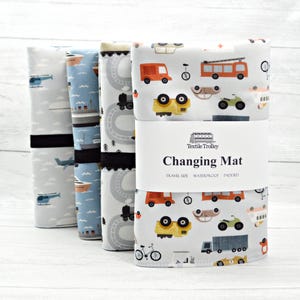 May include: A stack of travel-size, waterproof, padded changing mats with various patterns. Designs include vehicles, trains, and clouds. The front mat features a white band with the words "Changing Mat" and the brand name "Textile Trolley".