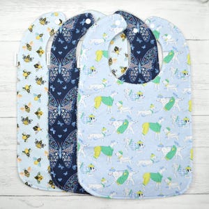May include: Three baby bibs featuring distinct patterns: one with yellow and black bees on light blue, one with dark blue butterflies, and one with white dogs wearing green and yellow sweaters on light blue. Each bib has a white snap closure.