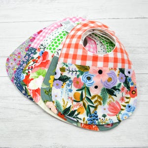 May include: A stack of five fabric baby bibs featuring various patterns including orange and white gingham, large colorful floral prints, green polka dots, and small purple and pink floral designs. Each bib has a rounded neck opening.