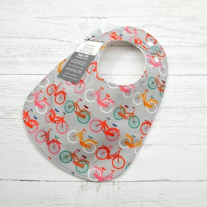 Puede incluir: Un babero gris para beb&eacute; con un estampado de bicicletas de colores. El babero tiene un forro blanco y un cierre de presi&oacute;n. El texto de la etiqueta dice "Textile Trends Baby Bib 2-piece set 2-3 years".