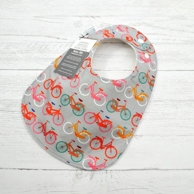 Puede incluir: Un babero gris para beb&eacute; con un estampado de bicicletas de colores. El babero tiene un forro blanco y un cierre de presi&oacute;n. El texto de la etiqueta dice "Textile Trends Baby Bib 2-piece set 2-3 years".