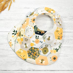 May include: A yellow and white baby bib with a honeycomb pattern and bee illustrations. The bib has a snap closure.