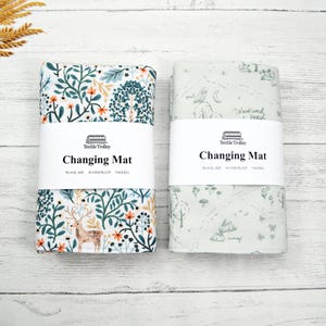 May include: Two rolled-up changing mats. One features a forest scene with a deer, trees, and floral elements. The other has a woodland map design with an owl, moon, and bunny. Each mat has a white band with the text "Changing Mat".