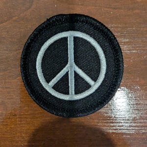 May include: A circular embroidered patch featuring a black background with a white peace symbol stitched in the center. The patch has a thick, black stitched border around the entire circumference, creating a defined edge for the circular design.