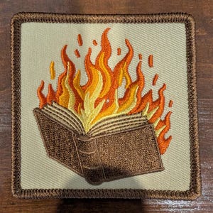 May include: A square embroidered patch with a tan background and a dark brown stitched border. The design features an open book with dark brown covers and pages, engulfed in vibrant yellow, orange, and red flames rising upward.