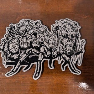 Pode incluir: Um emblema bordado com um design a preto e branco de uma figura esquelética com cabelo comprido e múltiplos olhos, carregando um grande pacote cheio de vários itens, incluindo cestos e recipientes, com um cajado segurado numa mão.