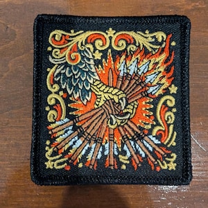May include: A square embroidered patch with a black background featuring a golden eagle talon clutching a bundle of arrows. The design includes vibrant orange and red flames, metallic gold scrollwork borders, and silver-tipped arrows.