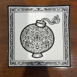 May include: A square embroidered patch featuring a black and white design of a round bomb with a lit fuse. The bomb is decorated with intricate floral patterns and is enclosed within a decorative rectangular border with a repeating scroll motif.