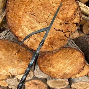 May include: A hand-forged black metal fire poker featuring a long, slender shaft with a distinct twisted design. The top end splits into a two-pronged fork shape, with one straight, pointed tine and one curved, tapered tine for moving logs.