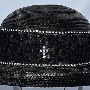 May include: A black straw hat featuring a wide brim and a rounded crown. The hat is decorated with a black lace band, two rows of clear rhinestones, and a small cross emblem made of clear rhinestones centered on the front of the band.
