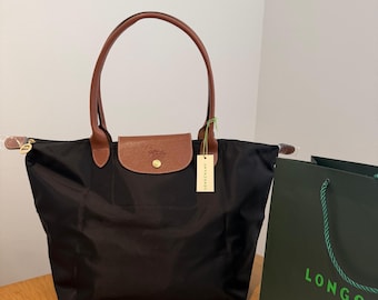 Puede incluir: Un bolso tote de nailon negro con asas de cuero marrón y una solapa de cuero marrón a juego con un cierre de botón a presión dorado. El bolso incluye una etiqueta colgante con la marca Longchamp y viene con una bolsa de compra verde oscuro con el texto LONGCHAMP PARIS.