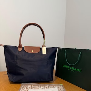 Op de afbeelding: Een marineblauwe Longchamp tote bag met bruine lederen handvatten en een klepsluiting met een goudkleurige drukknoop. Daarnaast staat een donkergroene Longchamp Paris-tas en een doorzichtige plastic zak met een herhalend logopatroon.