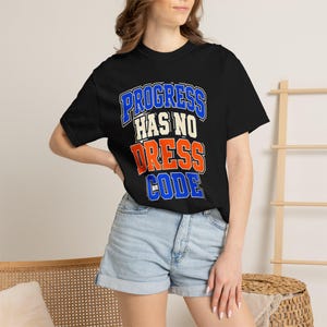 May include: A black short-sleeved t-shirt featuring the text "PROGRESS HAS NO DRESS CODE" printed in a bold, collegiate-style font. The word "PROGRESS" and "CODE" are in blue, "HAS NO" is in white, and "DRESS" is in orange with a white outline.