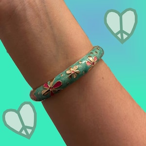 May include: A teal bangle bracelet featuring a repeating pattern of gold-toned flowers with pink and white enamel centers. The surface of the bracelet is smooth and glossy, showcasing a vibrant floral design against a solid teal background.