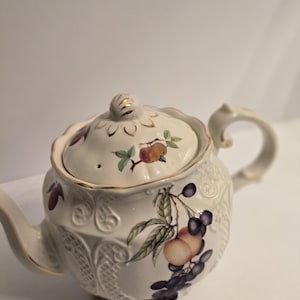 Puede incluir: Una tetera de cerámica color crema con una textura de celosía en relieve y un motivo de frutas pintadas que incluye uvas y melocotones. La tetera tiene una tapa a juego con un asa dorada, un pico curvo y detalles dorados en el borde.