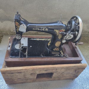 Könnte beinhalten: Eine schwarze Vintage-Nähmaschine der Marke Singer mit goldenen Blumenverzierungen und einem silbernen Handkurbelrad. Die Maschine ist auf einem dunklen Holzsockel montiert und teilweise in einer passenden Holzkiste untergebracht.