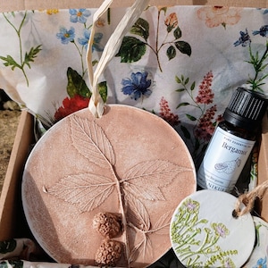 May include: A round terracotta-coloured clay ornament with a leaf and berry imprint, a smaller white circular ornament with green floral details, and a small amber glass bottle labelled Pure Essential Oil Bergamot in a box.