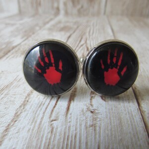 May include: Two circular cufflinks featuring a red handprint on a black background, set within a silver-toned metal frame. The handprint design is centrally positioned on each cufflink.