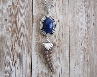 May include: A silver-toned pendant necklace featuring an oval, deep blue stone set in a decorative frame. Below, a carved, horn-shaped pendant hangs, creating a unique, eye-catching design. The necklace is displayed against a wooden background.