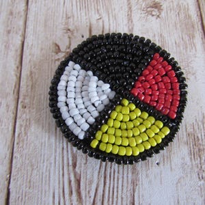 Può includere: Un disegno rotondo di perline con un bordo nero. Il disegno è diviso in quattro sezioni: bianco, rosso, giallo e nero. Le perline sono piccole e fitte, creando una superficie strutturata. Il disegno è su una superficie di legno chiaro.