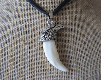 Pode incluir: Um pingente de cabeça de águia de prata com um amuleto branco em forma de dente pendurado em um cordão de couro preto.