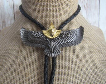 May include: A silver-toned bolo tie with a detailed eagle design and gold-coloured accents. The eagle's wings are spread, and the tie has a black braided cord. The bolo tie is displayed on a tan fabric background.