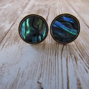 May include: Two round cufflinks with iridescent abalone shells set in gold-toned metal. The abalone shells display a mix of blue, green, and black colors. The cufflinks are displayed on a light-colored wooden surface.