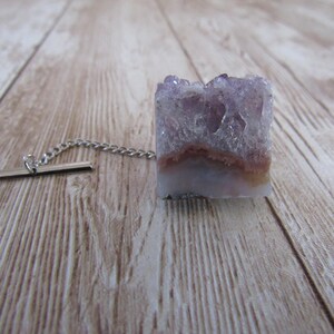 May include: A square amethyst tie tack with a silver chain and bar. The amethyst is a light purple color with a crystal structure and a brown band. The tie tack is on a light brown wooden surface.