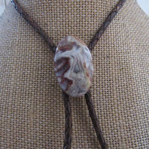 May include: A bolo tie featuring a large, oval-shaped stone pendant with a brown and cream marbled pattern. The pendant is suspended from a braided brown leather cord, displayed on a neutral-toned fabric.