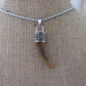 Puede incluir: Un collar con colgante que presenta una pieza marrón curvada en forma de cuerno, suspendida de una tapa de metal plateado con detalles intrincados de volutas. El colgante cuelga de una cadena de eslabones cuadrados de color plateado.