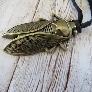 Può includere: Una collana con ciondolo a forma di cicala color bronzo con cordino nero. Il ciondolo a forma di insetto presenta dettagli sulle ali e sul corpo. La collana è esposta su una superficie di legno chiaro.