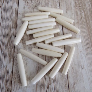 May include: A collection of off-white, cylindrical beads with rounded ends, scattered on a light-colored wooden surface. These beads are smooth and uniform in shape, suggesting they are for jewelry making or crafts.