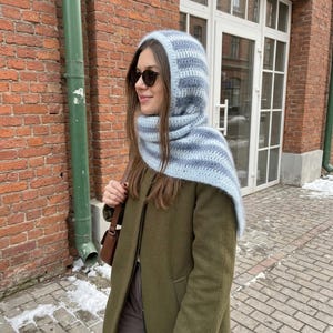 May include: A crochet balaclava hood in light blue and white horizontal stripes, worn over an olive green coat. The head covering features a soft, textured yarn construction that drapes around the neck and shoulders, paired with dark sunglasses.