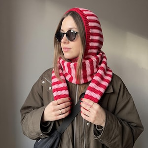 May include: A crochet balaclava hood and scarf combination featuring horizontal stripes in alternating shades of bright red and light pink. The accessory is worn over a dark olive green jacket and paired with round black sunglasses.