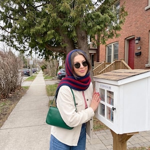 May include: A dark blue and red striped knit hood scarf, a cream-colored button-down jacket, blue jeans, and a solid green shoulder bag. The person wears dark round sunglasses and stands beside a white wooden Little Free Library box.
