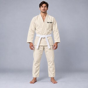 Puede incluir: Un uniforme de artes marciales de color beige que consta de una chaqueta de manga larga con cuello en V y pantalones a juego. La chaqueta presenta el texto "Your name" bordado en negro en el pecho izquierdo, sujeto con un cinturón blanco.
