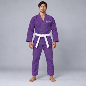 Puede incluir: Un kimono de artes marciales de color morado que incluye una chaqueta de manga larga con cuello en V y pantalones a juego. La chaqueta presenta el texto "Your name" bordado en blanco en el pecho. Un cinturón blanco ancho está atado a la cintura.