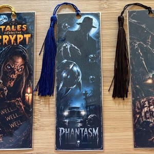 May include: Three rectangular bookmarks featuring horror movie themes. The first shows the Crypt Keeper with the text Tales from the Crypt and a yellow tassel. The second depicts Phantasm with a blue tassel. The third shows Jeepers Creepers with a black tassel.