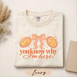 May include: An ivory t-shirt featuring a graphic print of a central orange bow flanked by two orange and yellow swirl designs. Below the graphics, the text reads, "you know why I'm here." The shirt has a Comfort Colors brand label at the neck.