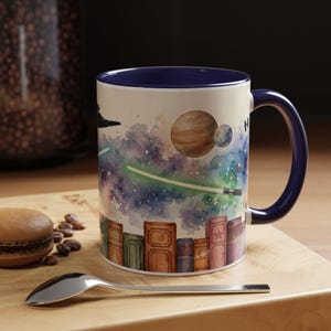 May include: A ceramic mug with a dark blue interior and handle, featuring a watercolour illustration of a row of books at the base, a starry nebula, planets, a spaceship, and a glowing green lightsaber blade extending across the white surface.
