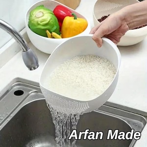 May include: A white plastic colander bowl with a perforated side for draining water, containing white rice being rinsed over a stainless steel sink. The text Arfan Made is visible in the lower right corner of the image near the water stream.