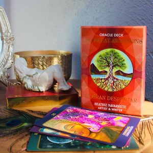 May include: A rectangular oracle deck box titled The Great Awakening El Gran Despertar by Beatriz Navarrete, featuring a central circular illustration of a tree with roots and a DNA strand. Several colorful oracle cards are fanned out in front.