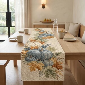 Puede incluir: Un camino de mesa acolchado con un estampado de calabazas azules y blancas rodeadas de hojas de otoño en tonos naranja, amarillo y verde. La tela tiene una superficie texturizada con costuras y está extendida sobre una mesa de madera.