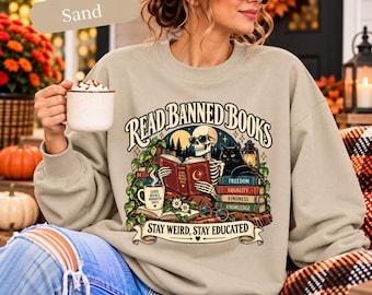 May include: A sand-colored crewneck sweatshirt featuring a graphic print of a skeleton reading a book with a black cat, a lantern, and a stack of books labeled Freedom, Equality, Kindness, and Knowledge. The text reads Read Banned Books.