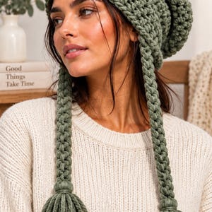 May include: A sage green crochet beanie featuring a textured, chunky knit pattern. The hat includes two long, braided side ties that hang down, each finished with a thick, matching green yarn tassel at the bottom end for a decorative finish.
