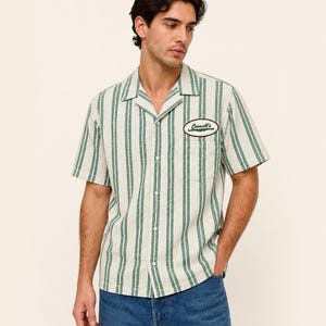 Peut inclure: Une chemise à manches courtes boutonnée avec un motif à rayures verticales crème et vert sur un tissu texturé. La chemise présente un col cubain, une poche poitrine unique et un écusson ovale sur la poitrine gauche avec le texte Cassell's.