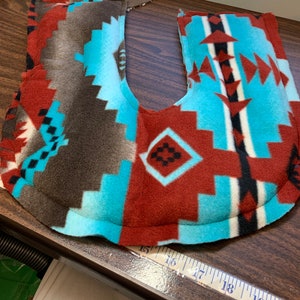 May include: A U-shaped neck pillow with a brown fleece backing and a colorful southwestern patterned fleece front. The pillow is on a wooden table with a measuring tape.