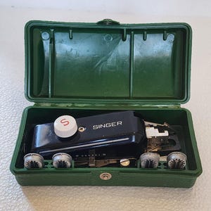 May include: A black Singer buttonholer attachment housed in a dark green plastic storage case. The device features a white dial with a red S, silver metal components, and four small circular metal bobbins arranged in the base of the case.